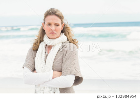 Woman gazing downward on sandy shoreline wearing knit poncho and scarf beside ocean waves 132984954