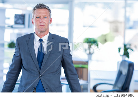 Middle-aged man standing in modern office near desk with computer monitor and potted plants 132984990