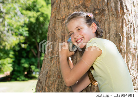 Female child leaning against tree trunk in grassy park listening on tin can telephone, copy space 132984991