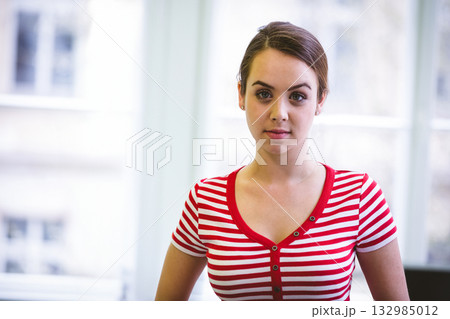 Woman standing facing camera by floor-to-ceiling windows in bright studio wearing striped shirt 132985012