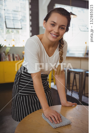 Female cafe server wearing striped apron leaning over table wiping with cloth in cafe by windows Female cafe server wearing striped apron leaning over table wiping with cloth in cafe by windows 132985015