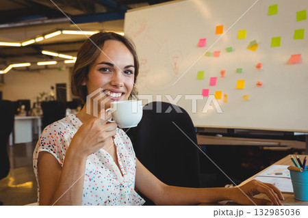 Professional woman sipping coffee from mug and smiling at office desk with whiteboard and pencils Professional woman sipping coffee from mug and smiling at office desk with whiteboard and pencils 132985036