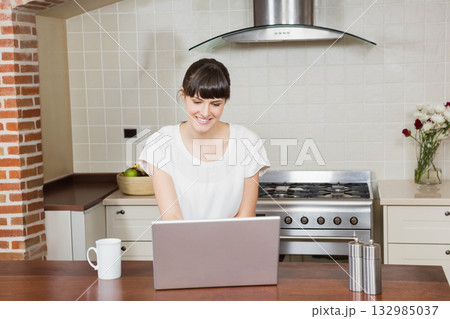 Woman sitting at kitchen island working on laptop while showcasing mug, fruit bowl 132985037