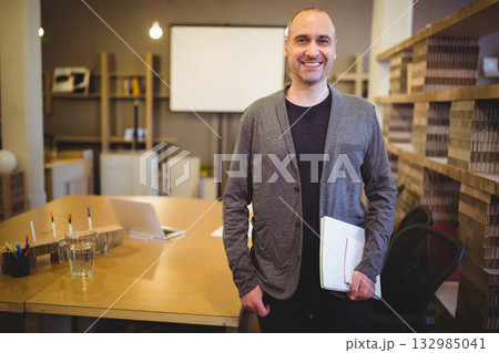 Man standing and smiling in modern office meeting area holding papers and notebook near laptop 132985041