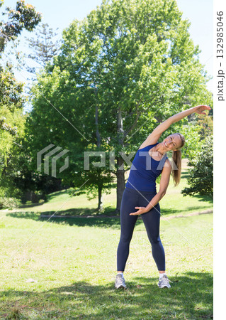 Woman performing side stretch on grassy park lawn in athletic wear and running shoes, copy space 132985046