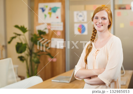 Red-haired woman in her 20s standing at office table with laptop and corkboard map, copy space Red-haired woman in her 20s standing at office table with laptop and corkboard map, copy space 132985053