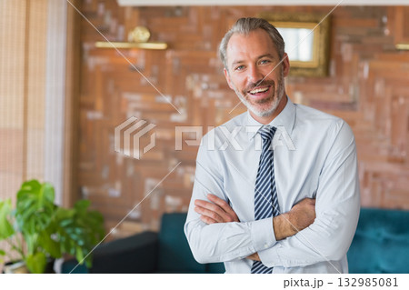 Businessman in shirt and tie standing arms folded in lounge with wood panels and teal sofa Businessman in shirt and tie standing arms folded in lounge with wood panels and teal sofa 132985081