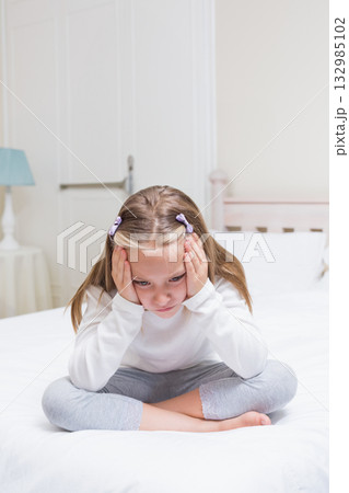 Female child sitting cross-legged pressing hands against head on bed in bedroom with table lamp Female child sitting cross-legged pressing hands against head on bed in bedroom with table lamp 132985102
