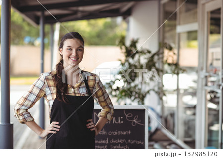 Female barista smiling while standing outside cafe under covered walkway with chalkboard menu sign 132985120