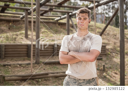 Male military trainee standing on wooden obstacle course wearing camo pants, muddy shirt and earbud 132985122