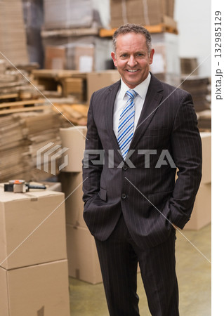 Middle-aged man wearing business suit standing in warehouse among boxes, pallets and tape dispenser Middle-aged man wearing business suit standing in warehouse among boxes, pallets and tape dispenser 132985129