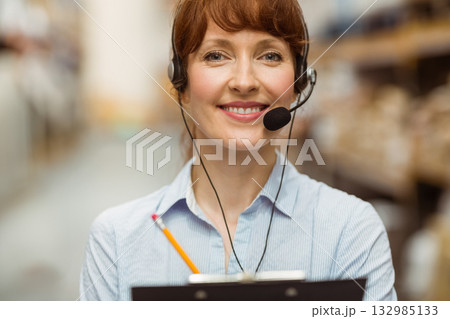Woman standing in warehouse aisle wearing headset holding clipboard near metal shelving and boxes Woman standing in warehouse aisle wearing headset holding clipboard near metal shelving and boxes 132985133