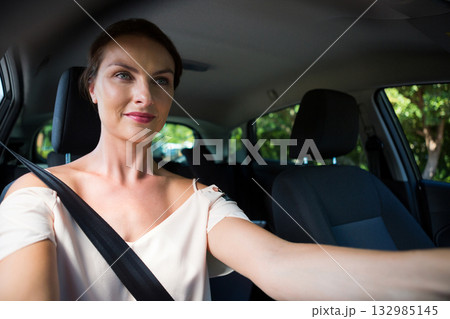 Female driver wearing seatbelt and gripping steering wheel while driving on sunny tree-lined road 132985145