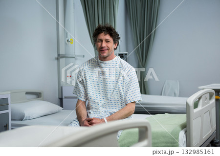 Mature male patient sitting on bed in patient room wearing hospital gown next to IV pole 132985161