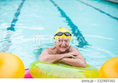 Senior woman leaning on inflatable ring in outdoor lap pool wearing yellow cap and black goggles 132985162