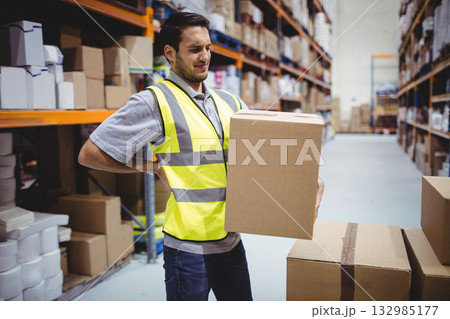 Male warehouse worker wearing safety vest lifting cardboard box amid metal racks in warehouse Male warehouse worker wearing safety vest lifting cardboard box amid metal racks in warehouse 132985177