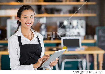 Female barista wearing apron, working with tablet at café by espresso machine, smiling, copy space 132985188