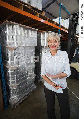 Senior female supervisor standing in warehouse by racks with forklift holding clipboard, copy space 132985190