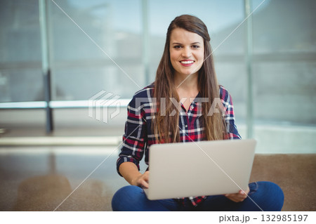 Woman wearing plaid shirt and jeans sitting on polished floor in study lounge using silver laptop Woman wearing plaid shirt and jeans sitting on polished floor in study lounge using silver laptop 132985197