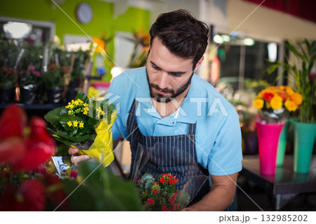 Male florist adjusting potted plant wrapped in yellow paper at workbench in shop with apron 132985202