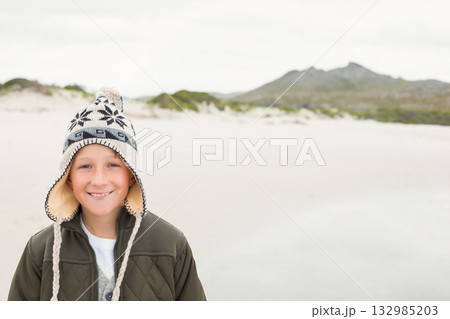Smiling child boy standing on sandy shore wearing olive green jacket and ear-flap hat, copy space Smiling child boy standing on sandy shore wearing olive green jacket and ear-flap hat, copy space 132985203