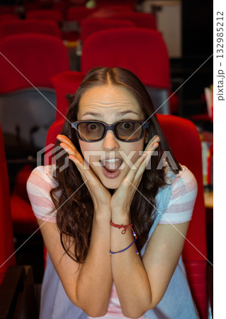 Female moviegoer wearing viewing glasses sitting in red cinema seats leaning forward in surprise Female moviegoer wearing viewing glasses sitting in red cinema seats leaning forward in surprise 132985212