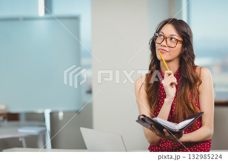 Businesswoman sitting in office holding planner and pencil beside laptop and whiteboard, copy space 132985224