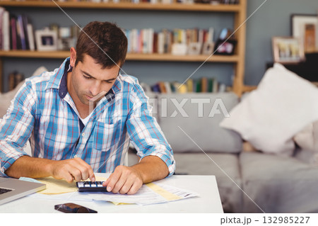 Man sitting at table in living room using calculator with laptop phone and documents, copy space 132985227