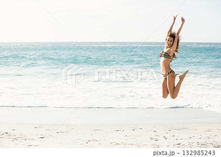 Woman in twenties jumping on sandy shoreline under midday sun wearing striped swimsuit, copy space Woman in twenties jumping on sandy shoreline under midday sun wearing striped swimsuit, copy space 132985243