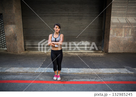 Japanese woman standing arms crossed on street by roll-up shutter, wearing sportswear and sweatband 132985245
