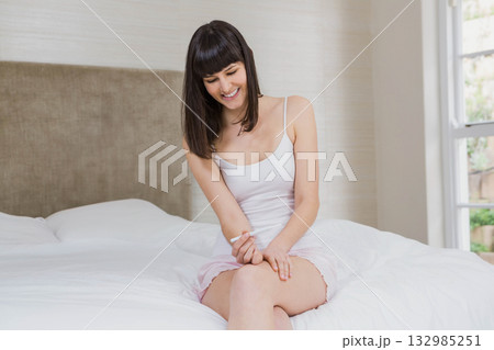 Woman sitting on neatly made bed near window in bedroom holding and smiling at white device 132985251