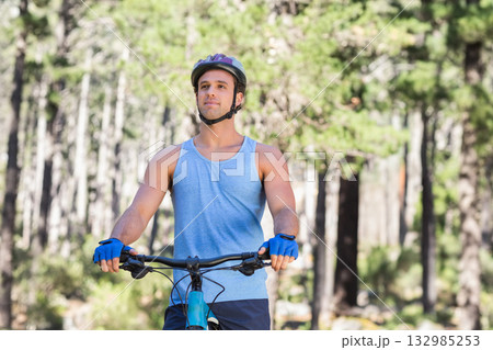 Man standing on pine forest trail wearing cycling helmet beside mountain bicycle under sunlight 132985253