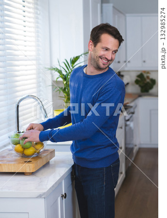 Man wearing blue sweater placing apples and lemons into glass bowl on cutting board in kitchen 132985274