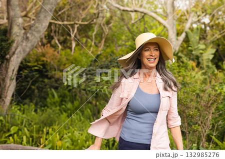 Senior woman walking along garden path wearing wide-brimmed straw sun hat and casual clothing Senior woman walking along garden path wearing wide-brimmed straw sun hat and casual clothing 132985276