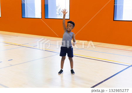 African American boy raising right arm while standing on multi-sport court under tall windows African American boy raising right arm while standing on multi-sport court under tall windows 132985277