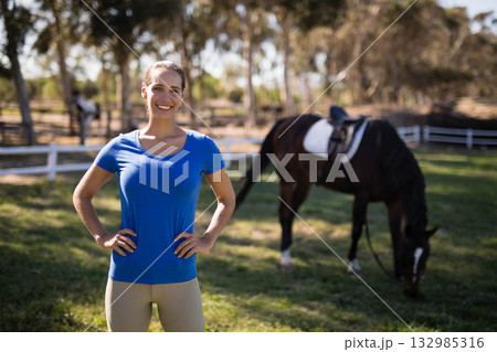 Smiling woman in riding gear standing in grassy paddock near white fence with saddled horse grazing Smiling woman in riding gear standing in grassy paddock near white fence with saddled horse grazing 132985316