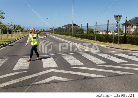Woman signaling traffic while wearing safety vest and crossing street at zebra crossing Woman signaling traffic while wearing safety vest and crossing street at zebra crossing 132985321