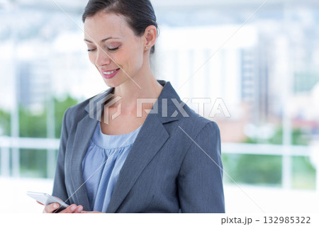 Businesswoman checking smartphone while standing at office windows overlooking city, copy space 132985322