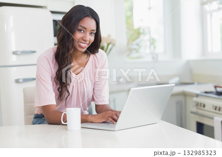 African American woman typing on silver laptop at white kitchen island near ceramic mug and flowers 132985323