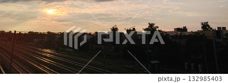 Cloudy evening sky with sun setting over railway lines and silhouetted buildings in the distance 132985403