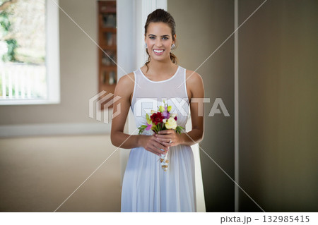 Woman standing in entryway holding colorful bouquet while wearing bridal gown by window blinds 132985415