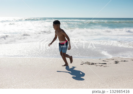 African American boy running along wet sand shoreline with ocean waves and striped swim trunks 132985416