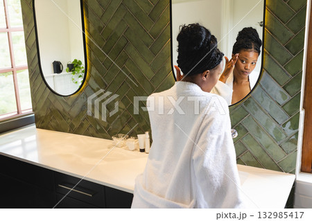 African American woman in white bathrobe applying facial serum at bathroom countertop with mirrors 132985417