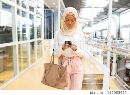 Woman walking airport walkway past glass windows and metal railings holding smartphone and tote bag 132985423