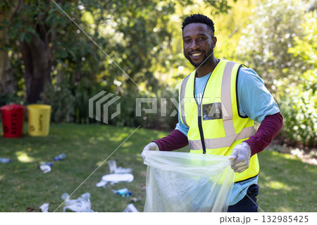 African American male volunteer picking litter on park lawn in vest holding trash bag, copy space 132985425