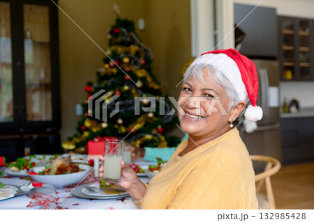 Senior woman wearing Santa hat sitting at dining table with gift and Christmas tree, copy space Senior woman wearing Santa hat sitting at dining table with gift and Christmas tree, copy space 132985428
