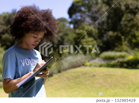 African American female volunteer jotting notes on clipboard with pen in sunlit park, copy space 132985429