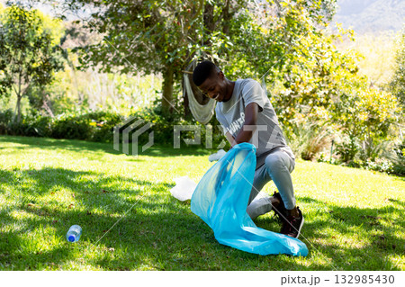African American man kneeling on grass placing water bottle into trash bag near hammock, copy space 132985430