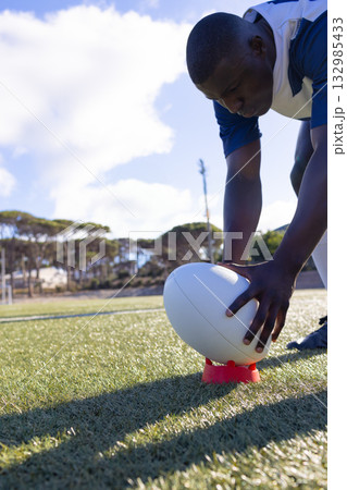 African American male athlete in jersey positioning rugby ball on kicking tee on pitch, copy space African American male athlete in jersey positioning rugby ball on kicking tee on pitch, copy space 132985433