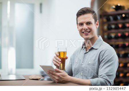Man wearing grey shirt sitting at bar counter holding beer glass, using tablet by wine rack 132985447